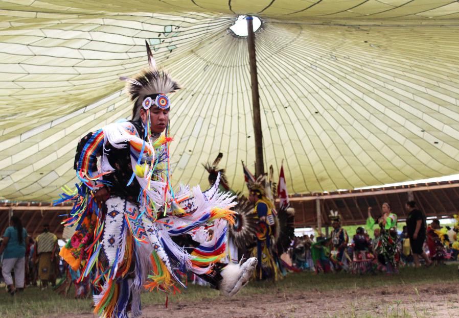 A First Nations dancer wears a colourful outfit and head piece and dances under a large tent at a pow wow.